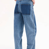 Back view of indigo denim patchwork pants on model wearing black t-shirt and black sandals, against white background.