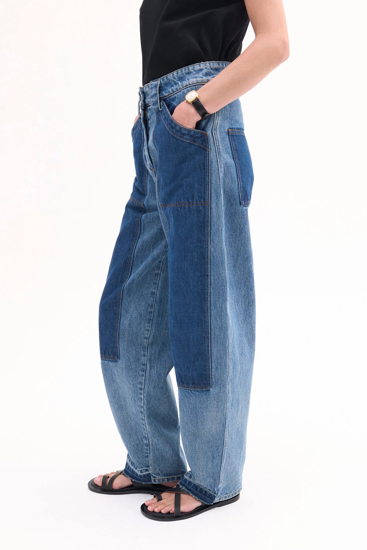 Side view of indigo denim patchwork pants on model wearing black t-shirt and black sandals, against white background.