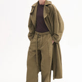 Front view of olive oversized trench coat on model wearing brown shirt, olive trousers and black pointed toe heels on white background.