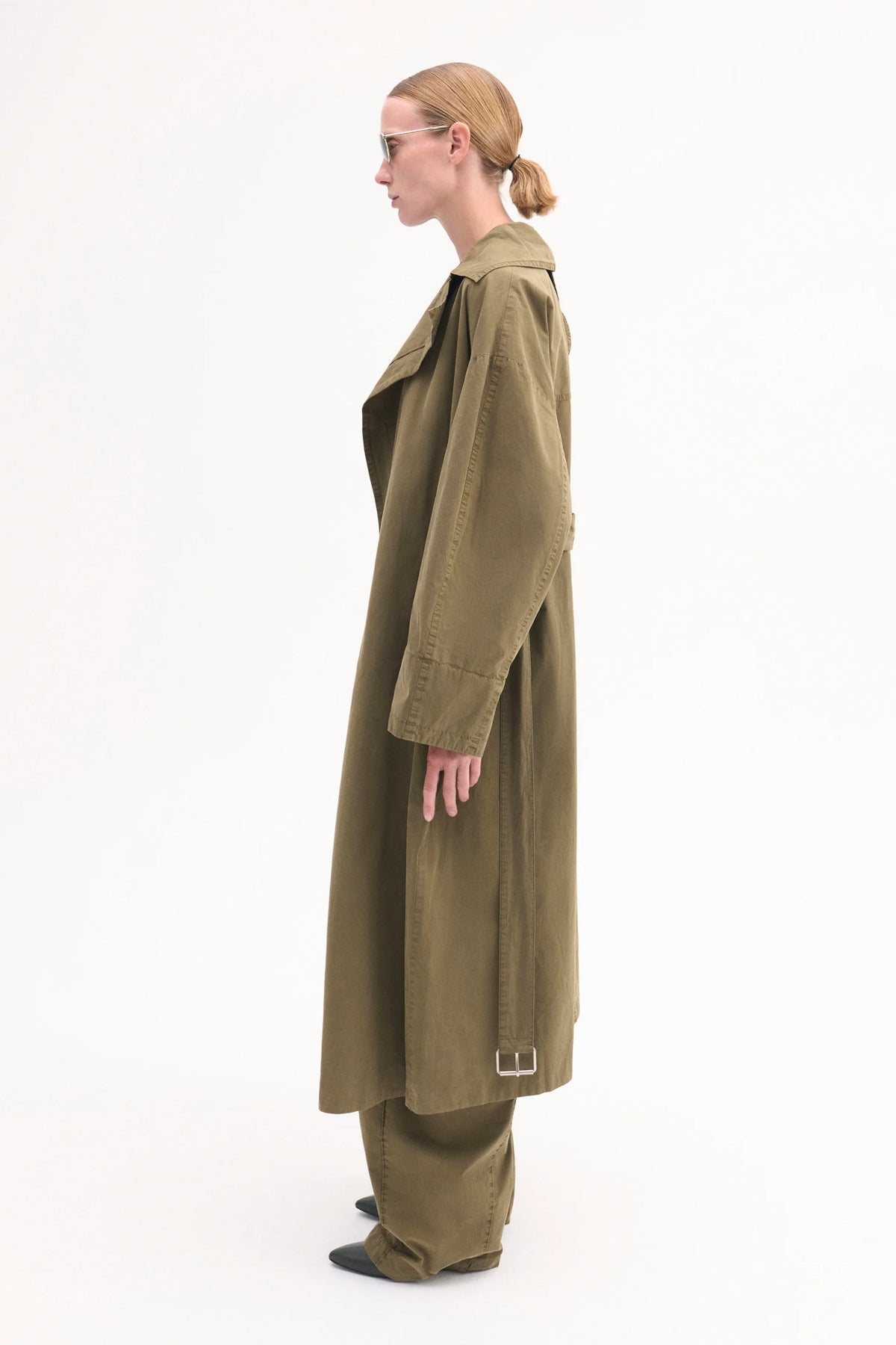 Side view of olive oversized trench coat mo model wearing brown shirt, olive trousers and black pointed toe heels on white background