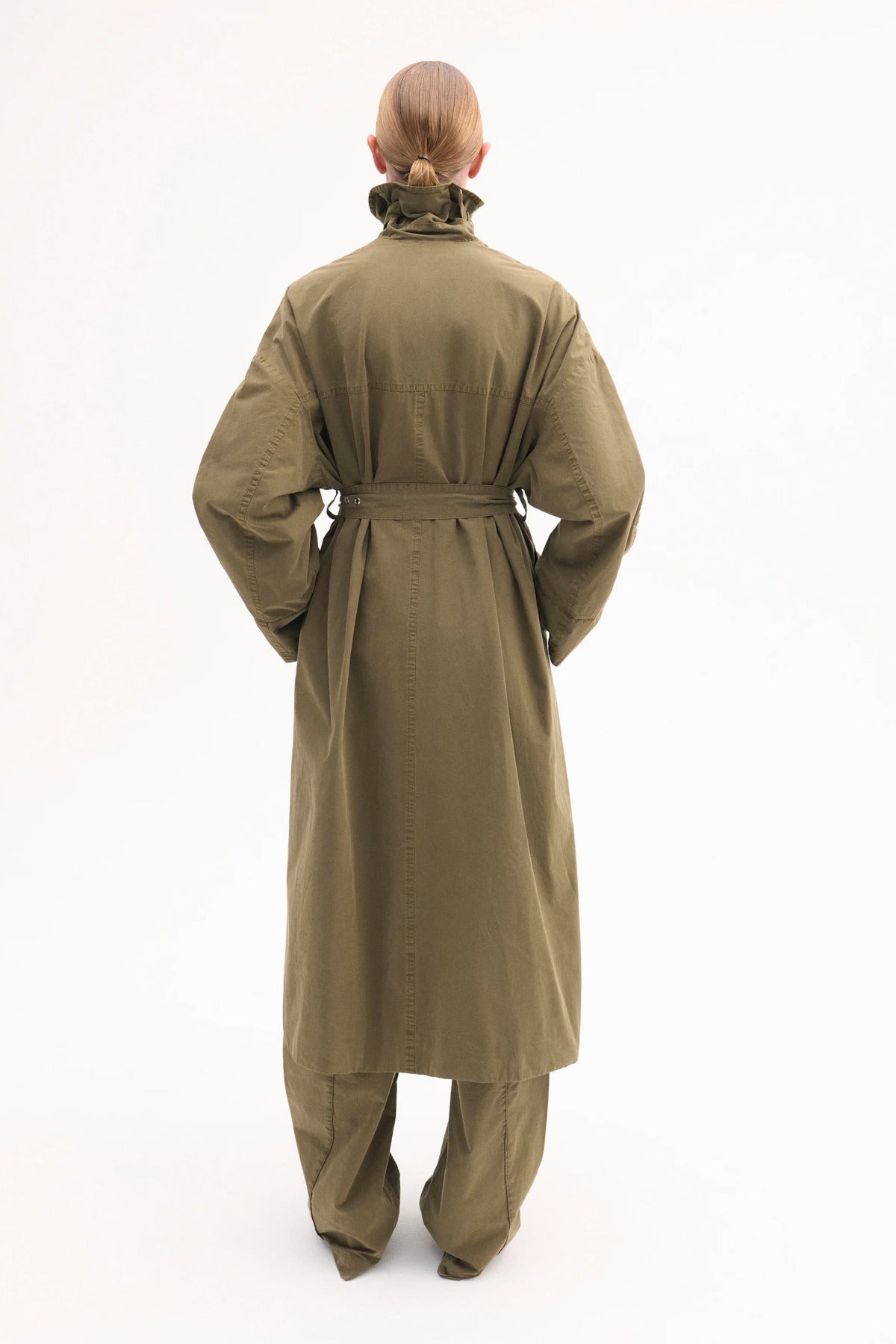 Back view of olive oversized trench coat on model wearing olive pants and black pointed toe heels on white background.
