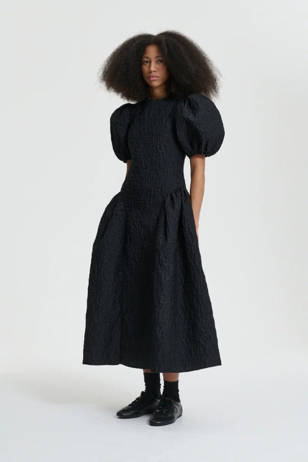 Front view of black bali dress on model wearing black sneakers and socks, against  a white background.