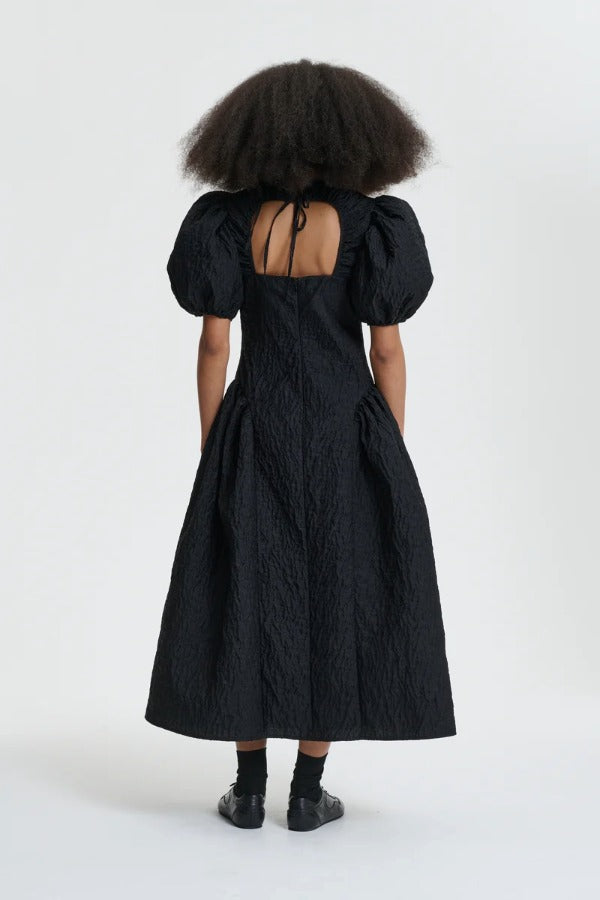 Back view of black bali dress on model wearing black sneakers and socks, against a white background.