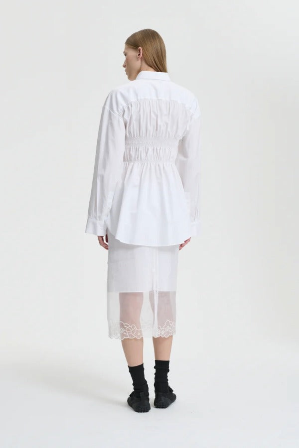 Back view of white braxton shirt on model wearing white slip and black sneakers, against a white background.