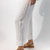 Side closeup view of pants with velvet ribbon on model wearing rice top and natural sandals, against a white background.