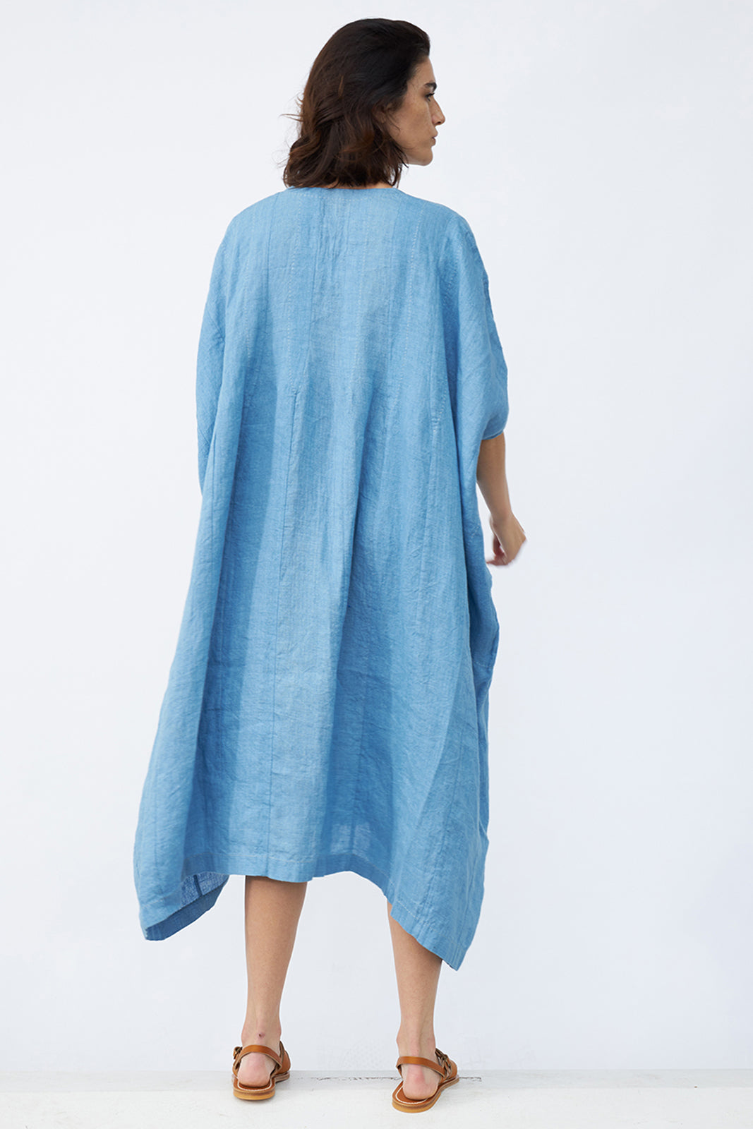 Back view of denim yoruba tunic on model wearing leather sandals, against a white background