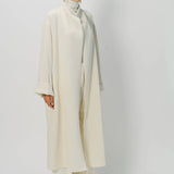Front slanted view of ivory long cardigan coat on model wearing white pants and turtleneck, against a white background.