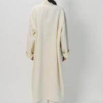 Back view of ivory long cardigan coat on model wearing white pants, against a white background.