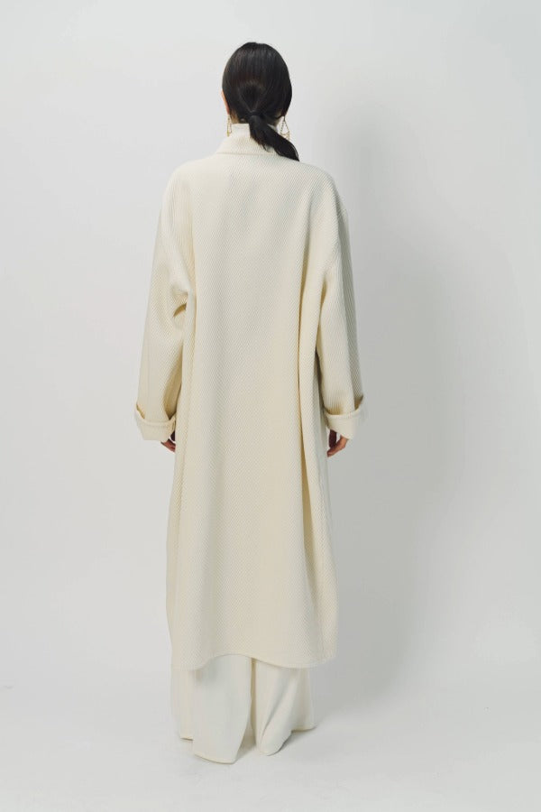 Back view of ivory long cardigan coat on model wearing white pants, against a white background.