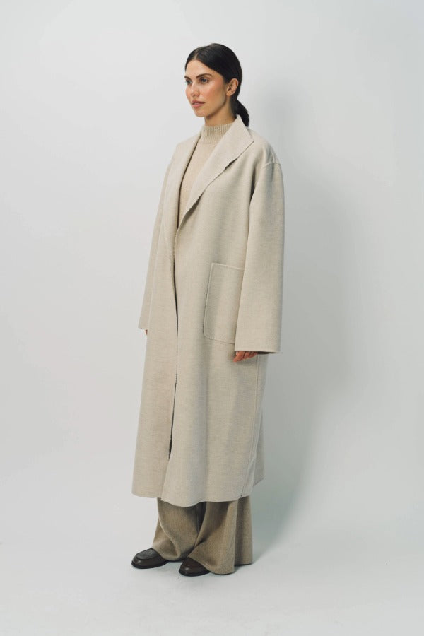 Side view of oatmeal cashmere patch pocket long coat on model wearing beige pants and oatmeal sweater, against a white background.