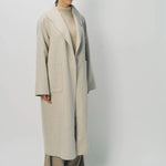 Front slanted view of oatmeal patch pocket long coat on model wearing beige pants and oatmeal sweater, against a white background.