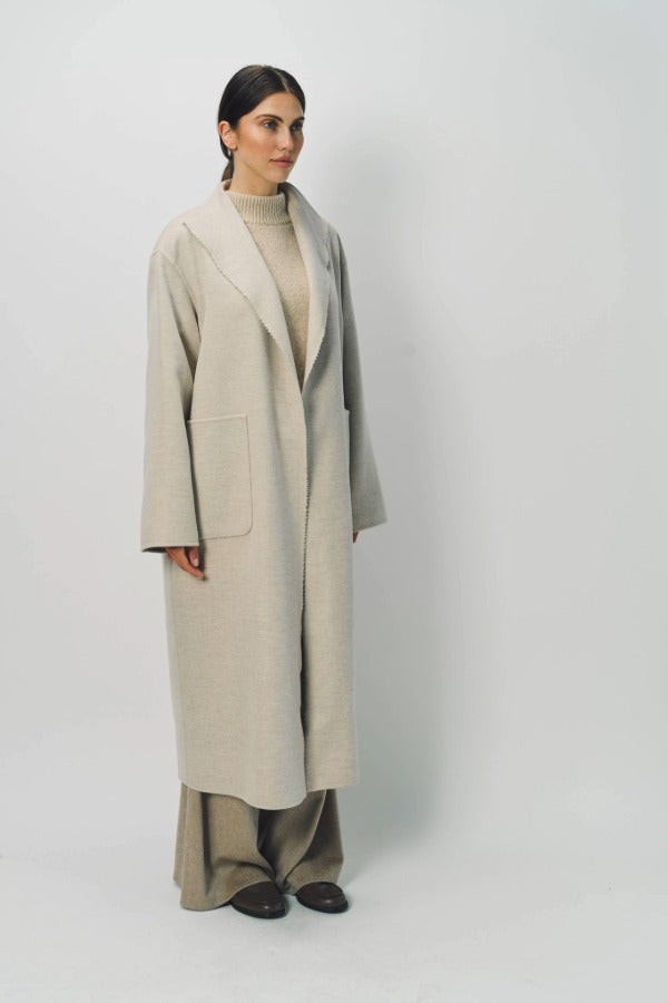 Front slanted view of oatmeal patch pocket long coat on model wearing beige pants and oatmeal sweater, against a white background.