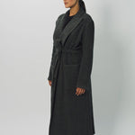 Side view of brown long cashmere jersey coat on model wearing black shirt and loafers, against a white background.