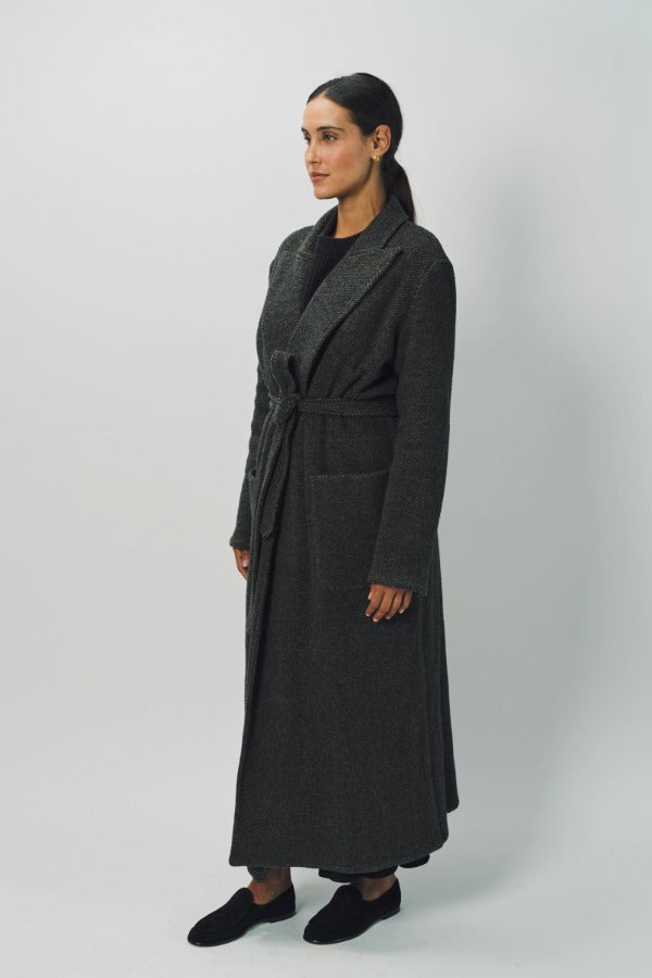 Side view of brown long cashmere jersey coat on model wearing black shirt and loafers, against a white background.