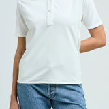 Front view of white daphne polo shirt on model wearing blue jeans, against a white background.