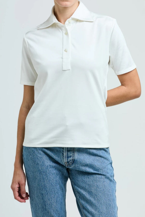 Front view of white daphne polo shirt on model wearing blue jeans, against a white background.