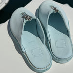Top-down view of a pair of light blue Guidi zip sandals resting on a white surface near the edge of a marina. The sandals feature central silver zippers and soft leather insoles stamped with “MADE IN ITALY.” The calm, dark water of the marina is visible in the background.