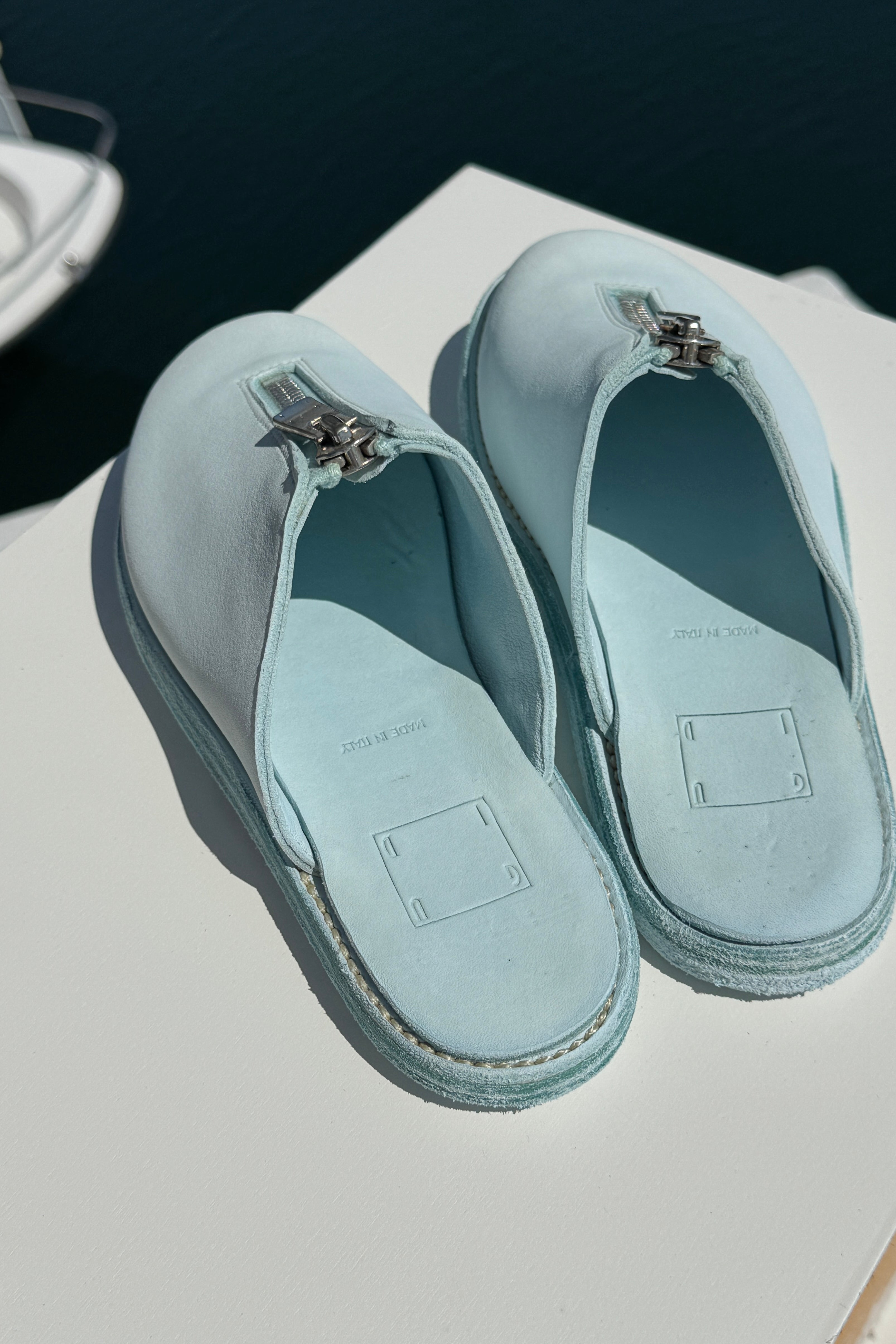 Top-down view of a pair of light blue Guidi zip sandals resting on a white surface near the edge of a marina. The sandals feature central silver zippers and soft leather insoles stamped with “MADE IN ITALY.” The calm, dark water of the marina is visible in the background.