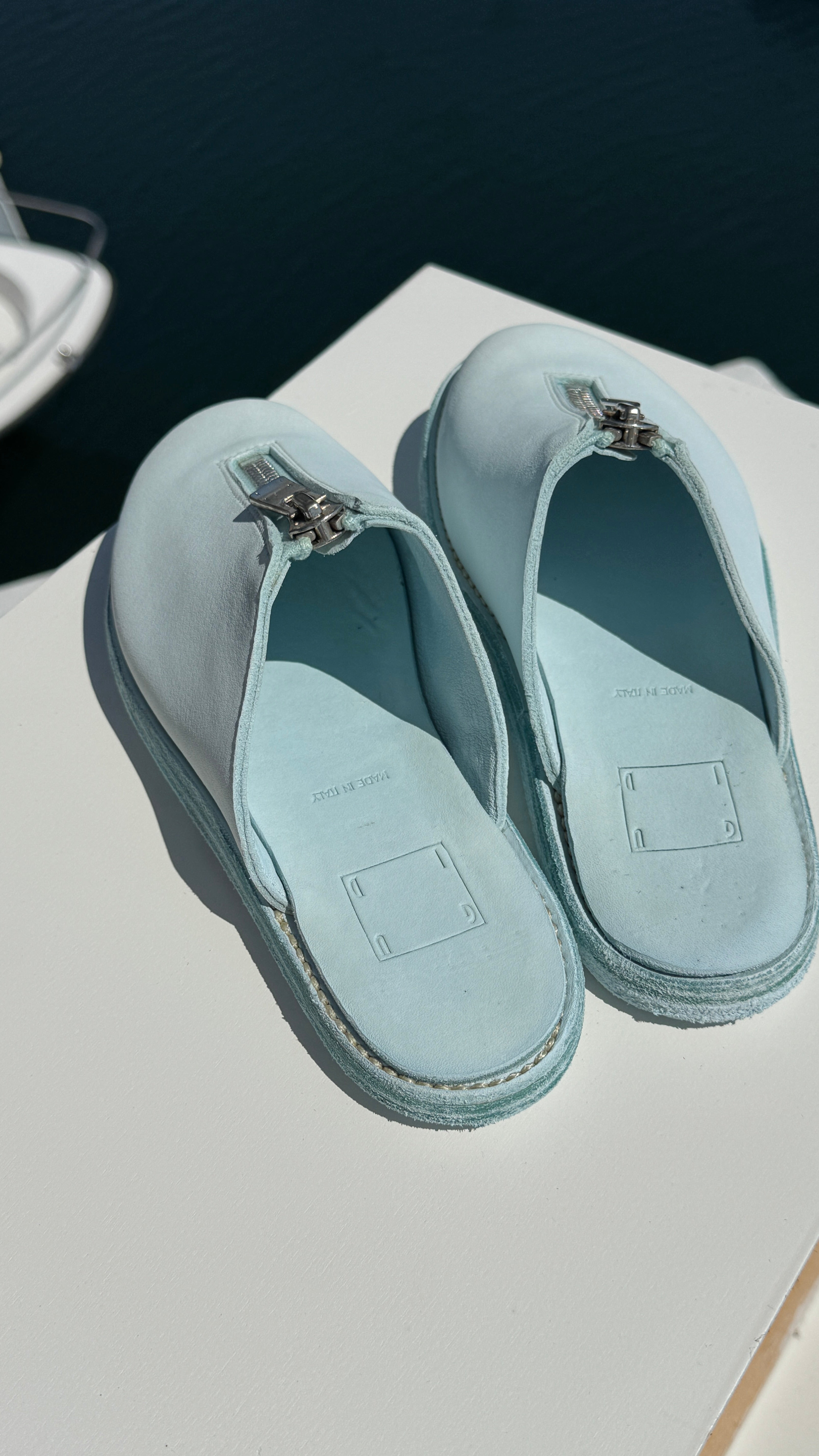 Top-down view of a pair of light blue Guidi zip sandals resting on a white surface near the edge of a marina. The sandals feature central silver zippers and soft leather insoles stamped with “MADE IN ITALY.” The calm, dark water of the marina is visible in the background.