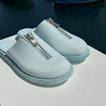 A pair of light blue Guidi zip sandals displayed on a white surface with a marina in the background. The sandals feature a central silver zipper and a textured sole, with yachts and boats visible in the blurred water behind them.