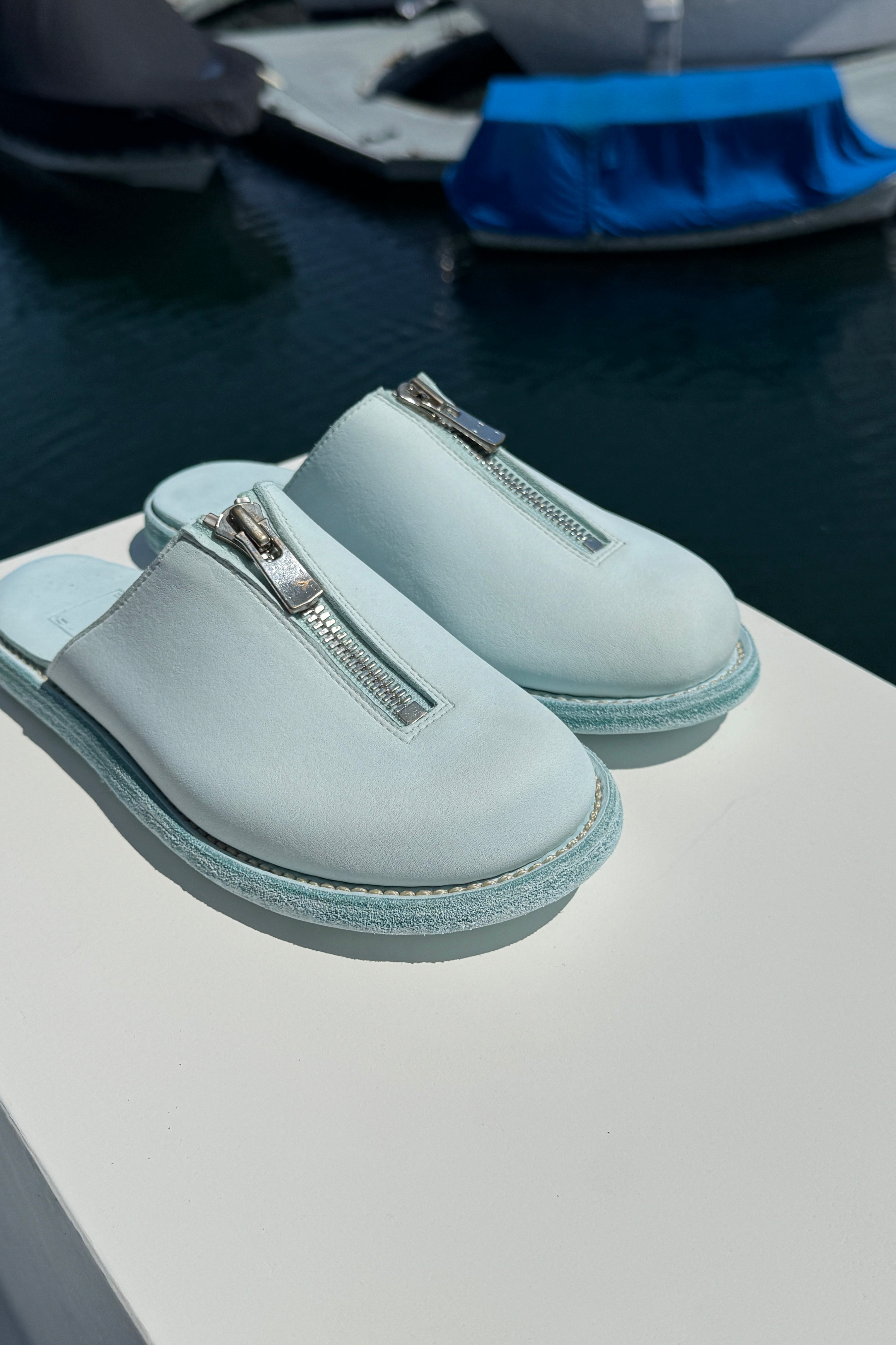A pair of light blue Guidi zip sandals displayed on a white surface with a marina in the background. The sandals feature a central silver zipper and a textured sole, with yachts and boats visible in the blurred water behind them.