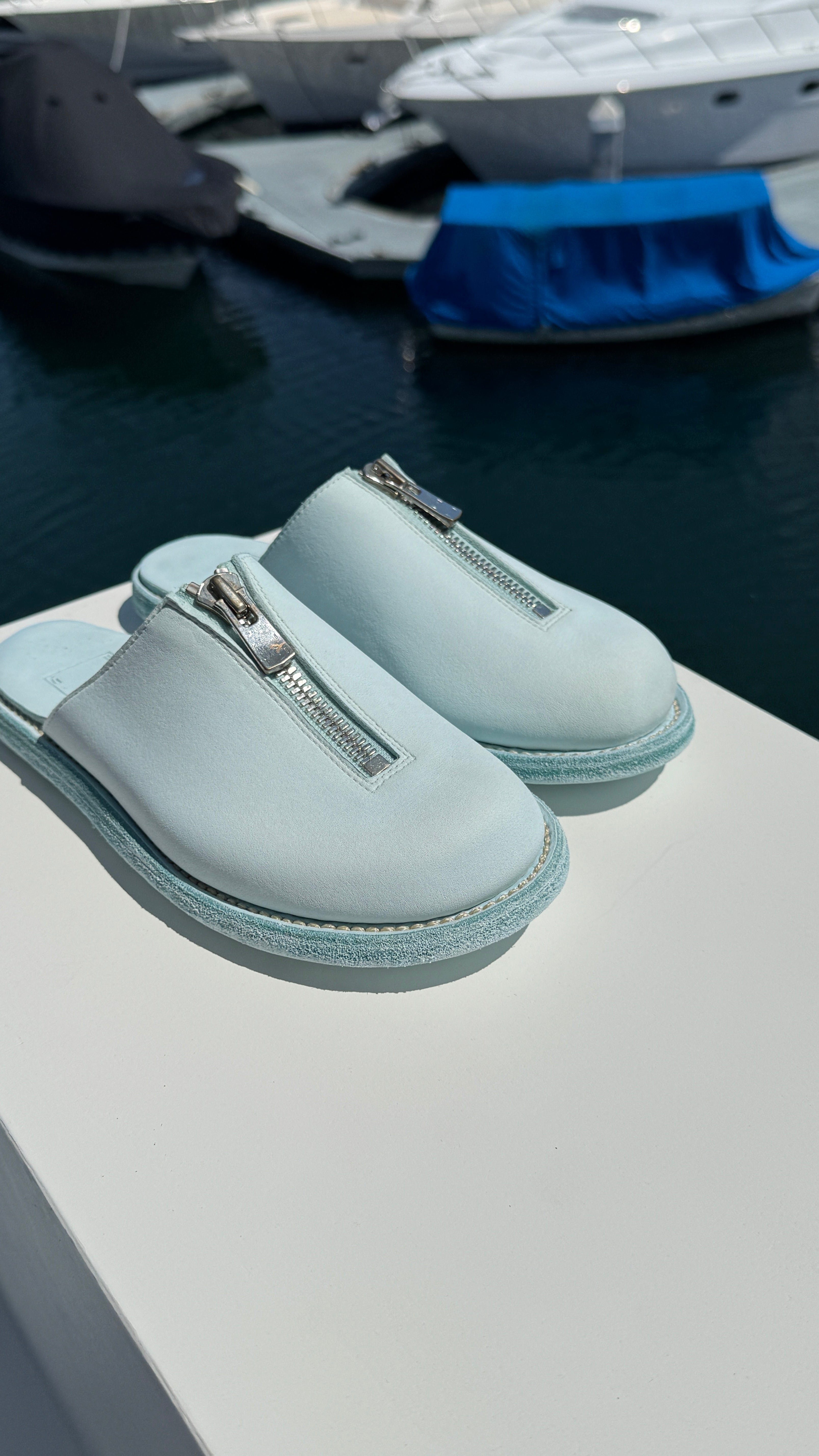 A pair of light blue Guidi zip sandals displayed on a white surface with a marina in the background. The sandals feature a central silver zipper and a textured sole, with yachts and boats visible in the blurred water behind them.