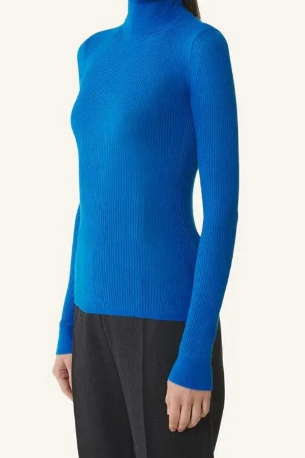 Side view of electric blue turtleneck jumper on model wearing grey pants, against a white background.
