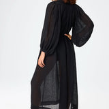 Slanted back view of black maxi poet dress on brown-haired model wearing brown sandals, against a white background.