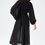 Back view of black maxi poet dress on brown-haired model wearing brown sandals against a white background.