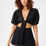 Front view of black pouf tie blouse on brown haired model wearing black shorts, against a white background.