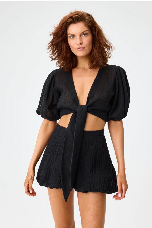 Front view of black pouf tie blouse on brown haired model wearing black shorts, against a white background.