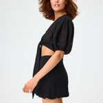 Side view of black pouf tie blouse on brown-haired model wearing black shorts, against a white background.