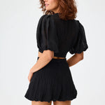 Back view of black pouf tie blouse on brown-haired model wearing black shorts against a white background.