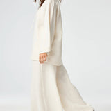 Side view of white low waist wide leg pant on brown-haired model wearing a white top and brown sandals, against a white background.
