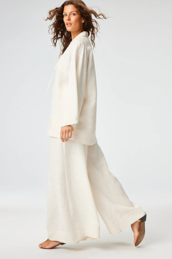 Side view of white low waist wide leg pant on brown-haired model wearing a white top and brown sandals, against a white background.