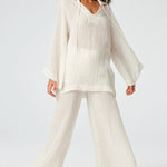 Front view of white tunic top on brown-haired model wearing white pants and silver sandals, against a white background.