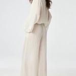 Side view of white tunic top on brown-haired model wearing white pants and silver sandals, against a white background.