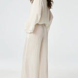 Side view of white tunic top on brown-haired model wearing white pants and silver sandals, against a white background.