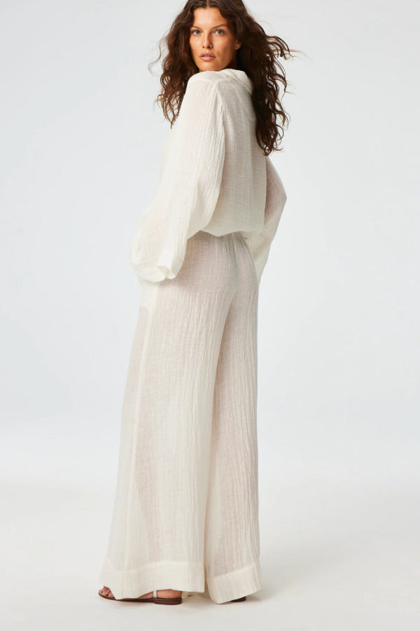 Side view of white tunic top on brown-haired model wearing white pants and silver sandals, against a white background.