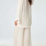 Back view of white tunic top on brown-haired model wearing white pants and silver sandals, against a white background.