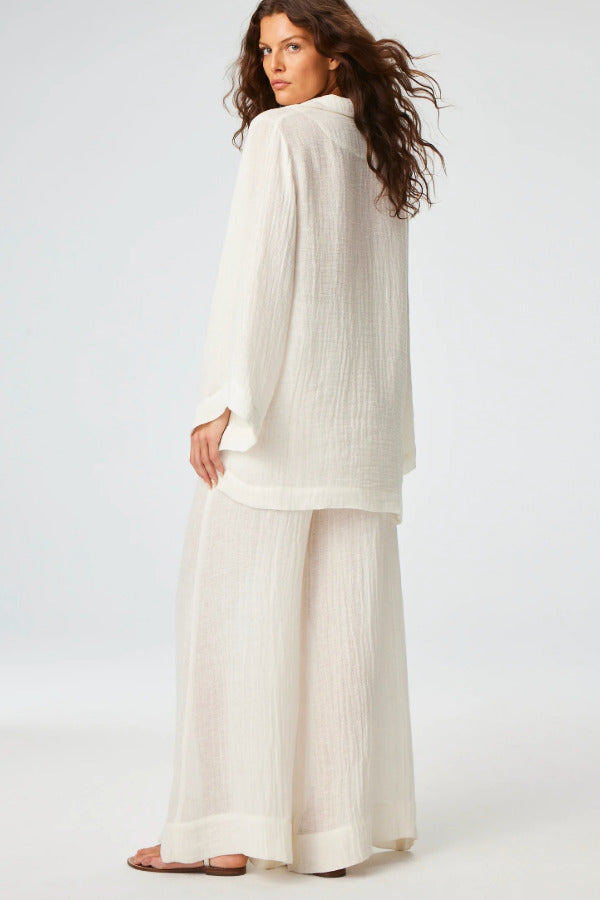 Back view of white tunic top on brown-haired model wearing white pants and silver sandals, against a white background.