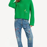 Front view of denim embroidered scribble jean on model wearing green sweater and pink slides, against a white background.