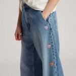 Side closeup view of mid blue embroidered straight leg jean on model wearing white t-shirt, against a white background.