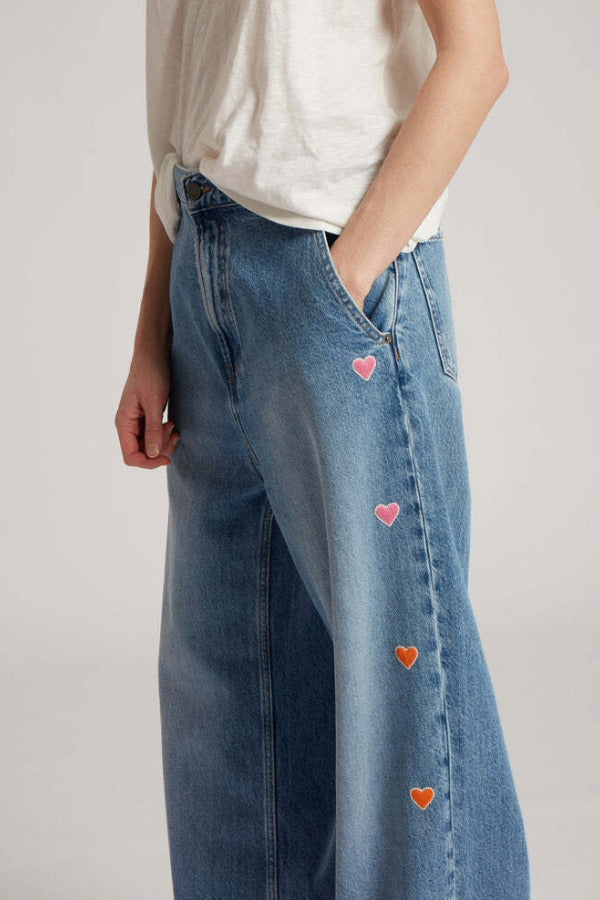 Side closeup view of mid blue embroidered straight leg jean on model wearing white t-shirt, against a white background.