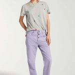Front view of lilac garment dyed jeans on model wearing grey shirt with hearts and white sneakers, against white background.