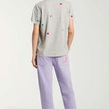 Back view of lilac garment dyed jeans on model wearing grey shirt with hearts and white sneakers, against a white background.