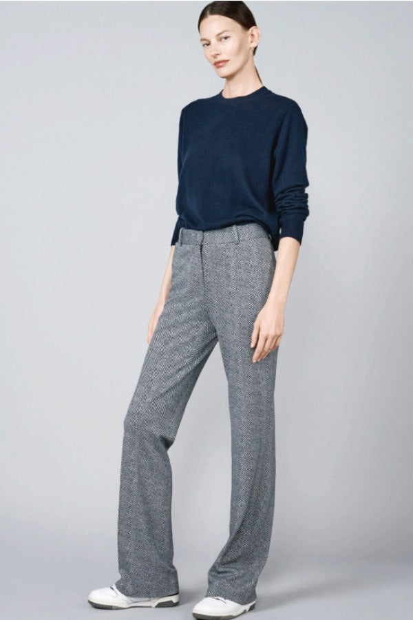 Front slanted view of navy blue herringbone paris pant on model wearing navy top and sneakers, against a grey background.