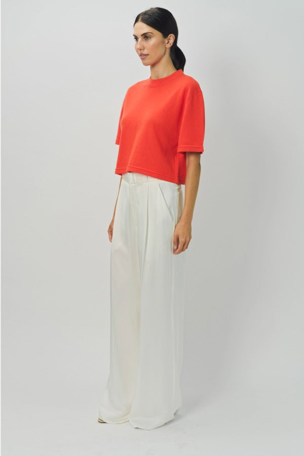 Side view of orange cashmere cropped t-shirt on model wearing white pants, against a white background.