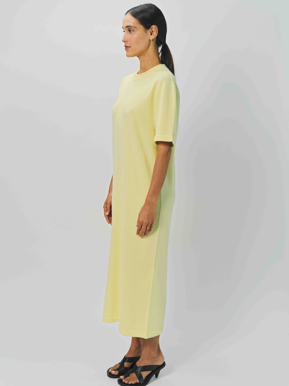 Side view of yellow cashmere long t-shirt dress on model wearing sandals, against a white background.