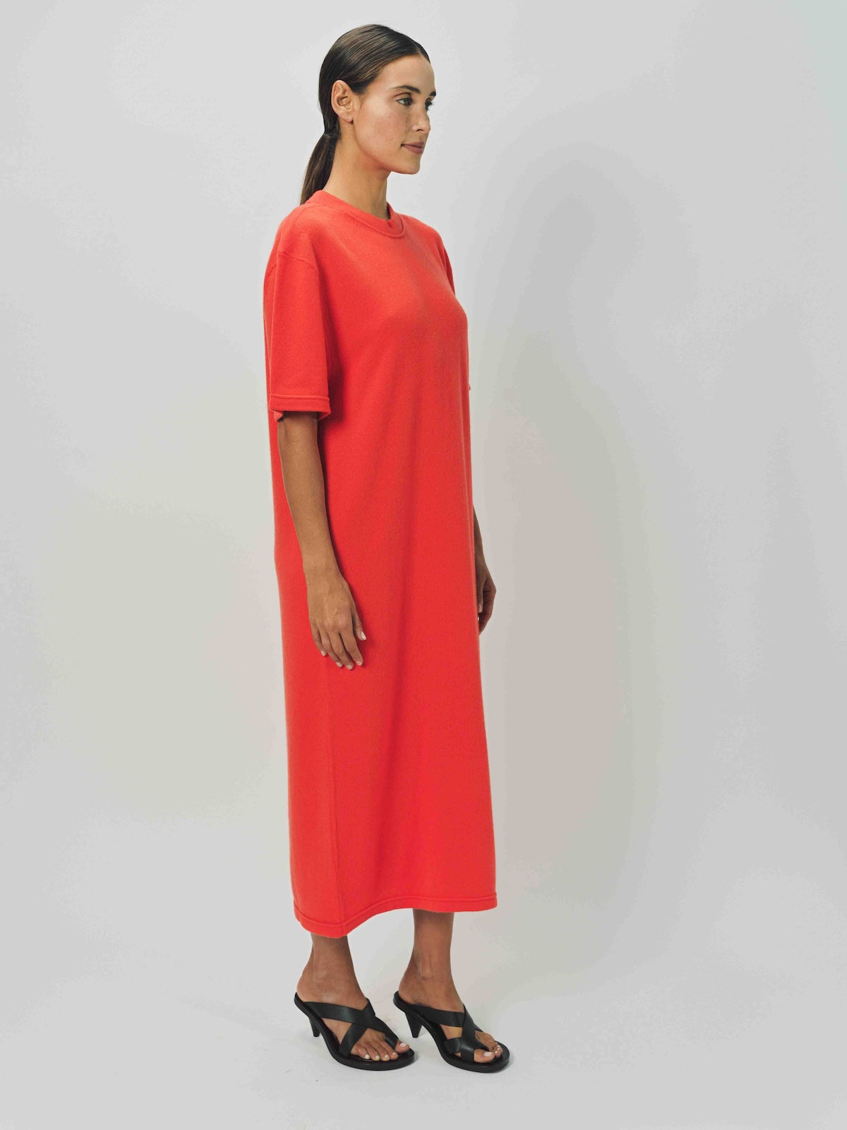 Side view of orange cashmere long t-shirt dress on model wearing sandals, against a  white background.